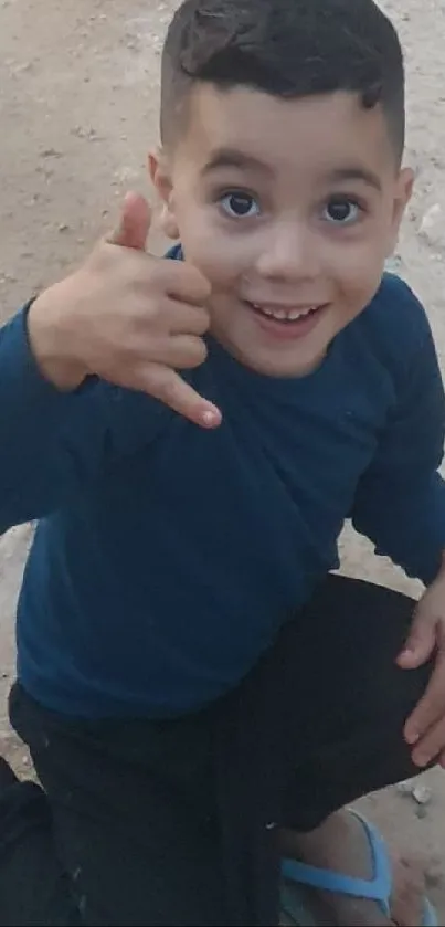 A smiling young boy kneels on sandy ground with a blue shirt.