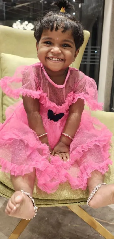 Smiling child in a pink dress sitting on a green chair indoors.