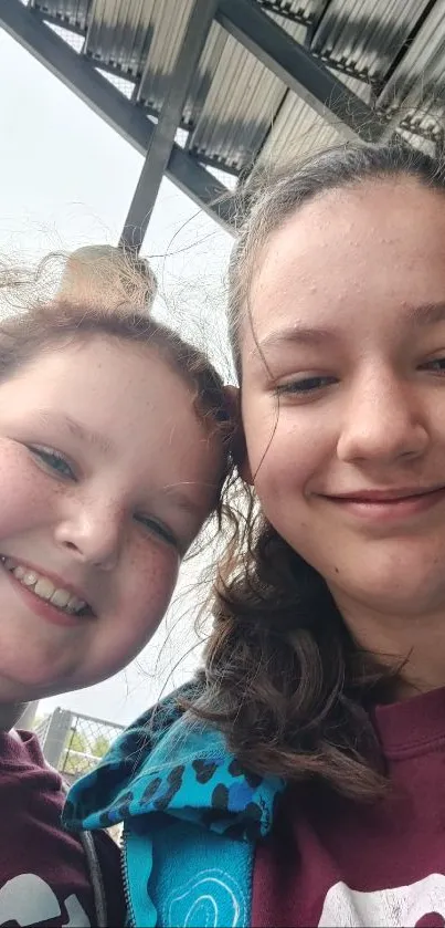 Two girls smiling under a metal roof, wearing casual clothing.