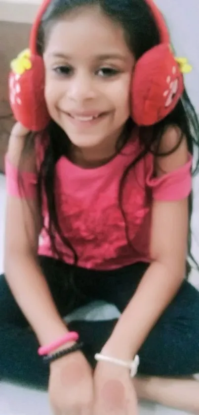 Young girl smiling in pink top and red earmuffs, sitting indoors.