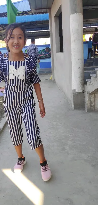 Girl in striped jumpsuit smiling outdoors near stairs.