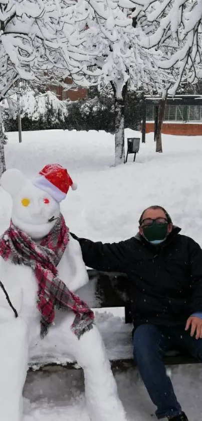 Snowman with a red hat and scarf next to a seated person on a snowy day.