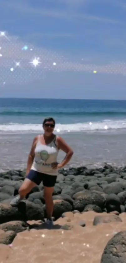 Woman on rocky beach with sparkling ocean under clear sky.