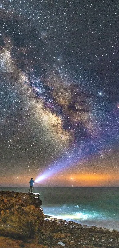 Starry night sky over ocean, person with flashlight on rocky shore.