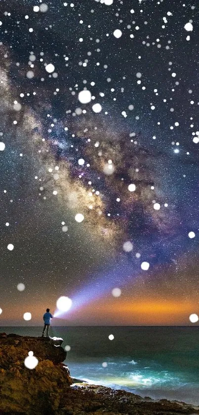 A lone figure shines a light towards a starry night sky above the ocean.