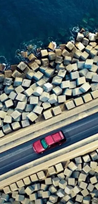 Aerial view of a red car on a coastal road with blue ocean and geometric rocks.