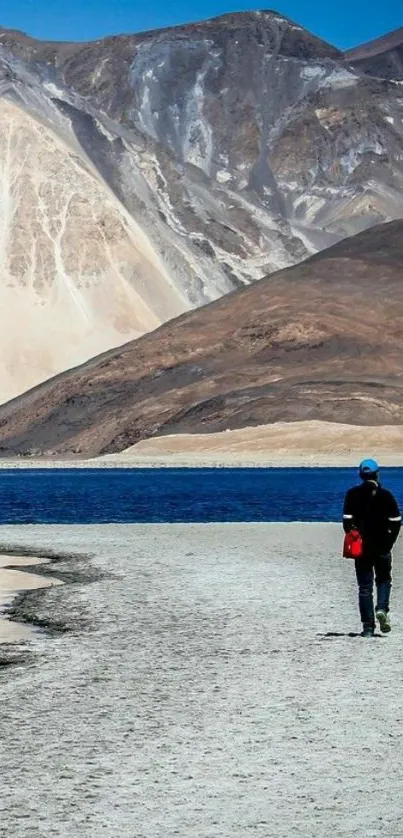 Person walking by blue lake against mountain backdrop.