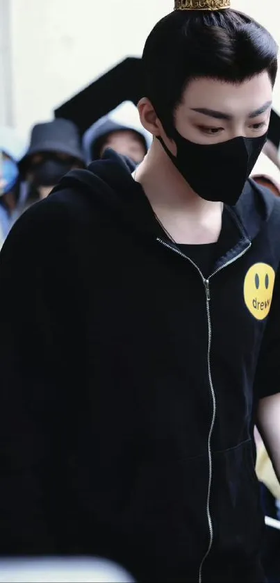 Person in a black hoodie with a yellow smiley design, wearing a black mask.