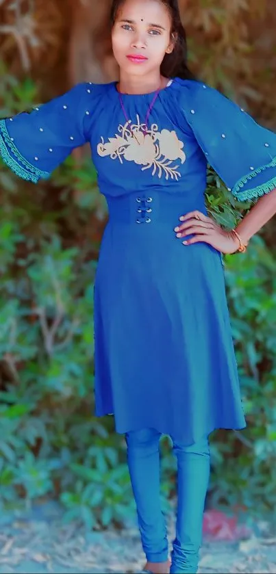 Person in a vibrant blue dress stands against a green leafy background.