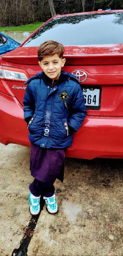 Boy in blue jacket leaning on a red car.