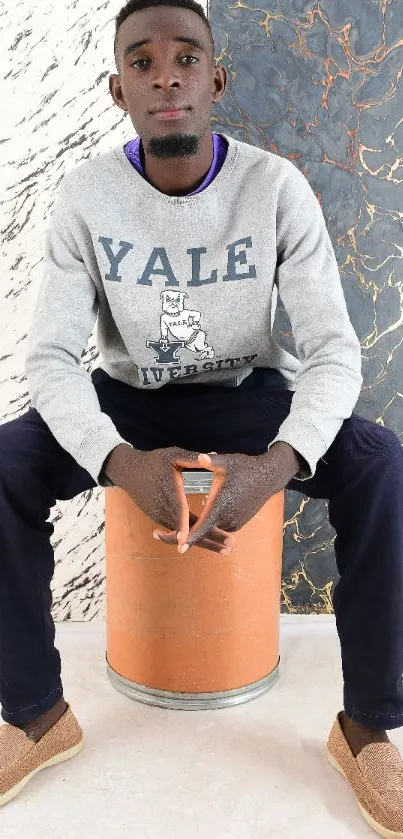 Stylish person in casual attire seated against textured wall.