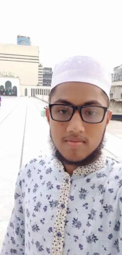 Person in traditional attire with city backdrop, wearing glasses and patterned shirt.