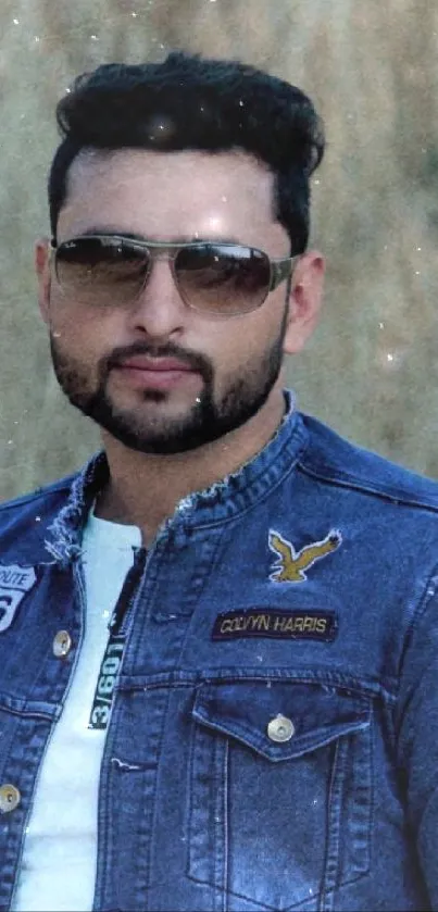 Man in denim jacket and sunglasses stands outdoors against a textured background.