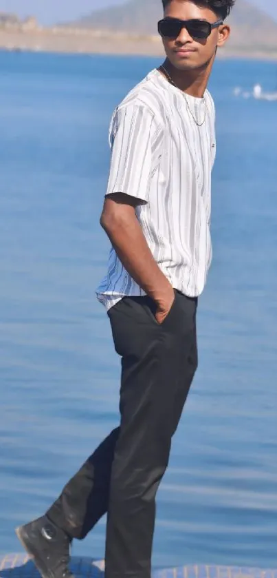 Man in stylish outfit walking by a lake with mountains in background.