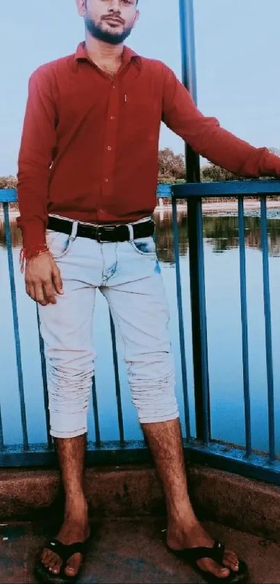 Man in red shirt and white jeans by a lake, leaning on railing.