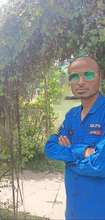 Man in a blue jumpsuit standing confidently in a lush garden setting.