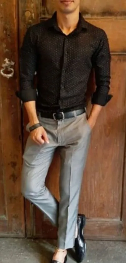 Man in black shirt and gray trousers leaning on wooden door.