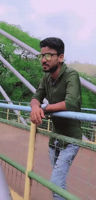 Man in green shirt leaning on bridge with lush greenery background.