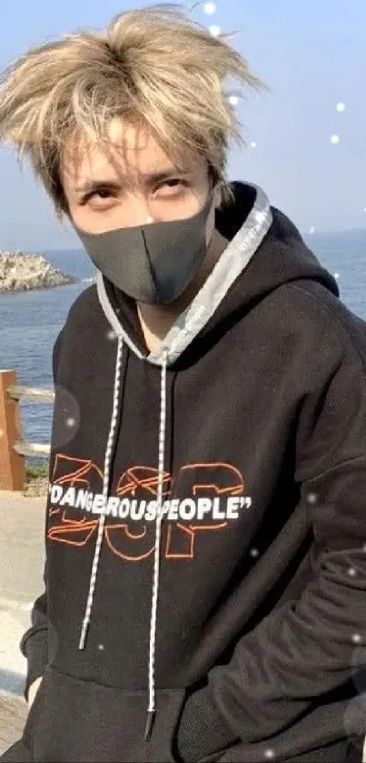 Young person in a black hoodie with mask by the sea, stylish and relaxed.