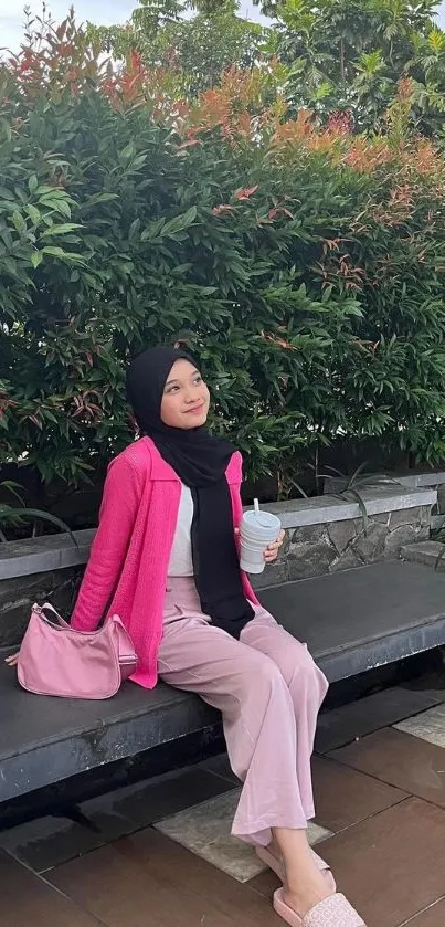 Woman in pink jacket sitting outdoors amid greenery.