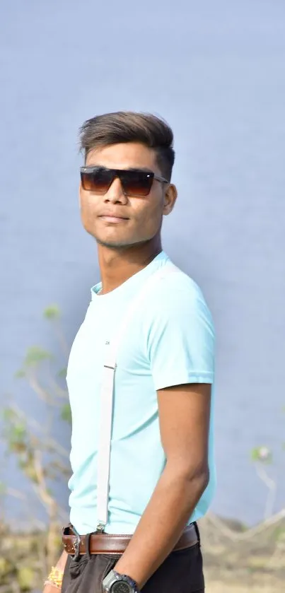 Stylish young man wearing sunglasses by a lake, dressed in a light blue shirt.