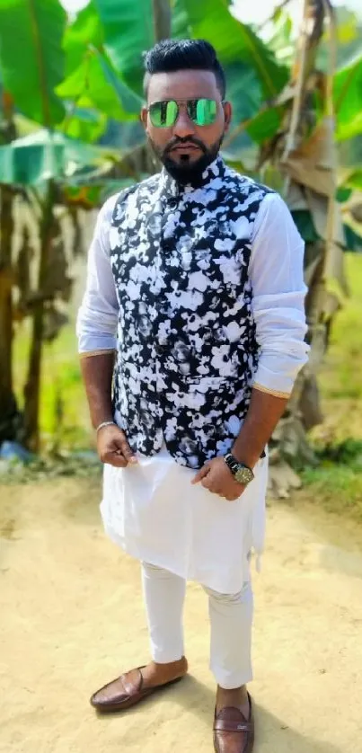 A stylish man poses outdoors, dressed in black and white, with vibrant green surroundings.