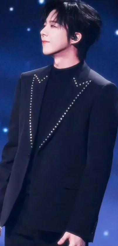 Performer in black suit on stage with blue background, holding microphone.