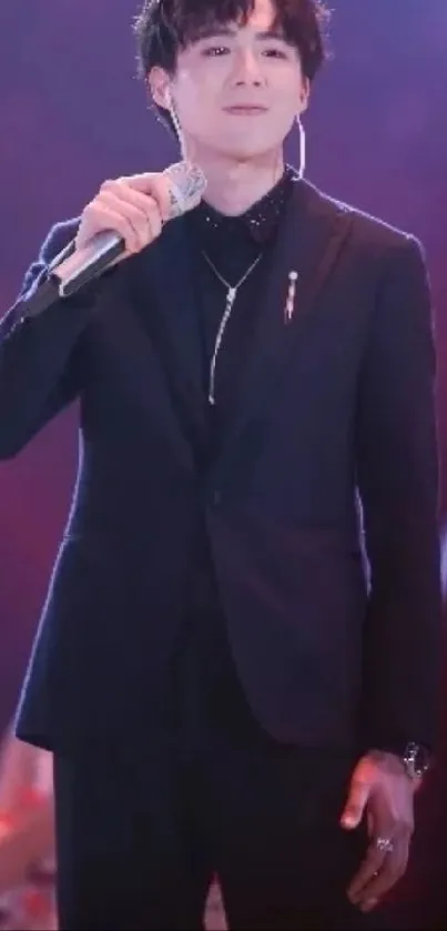 Confident performer on stage in a stylish black suit, set against a dark purple backdrop.