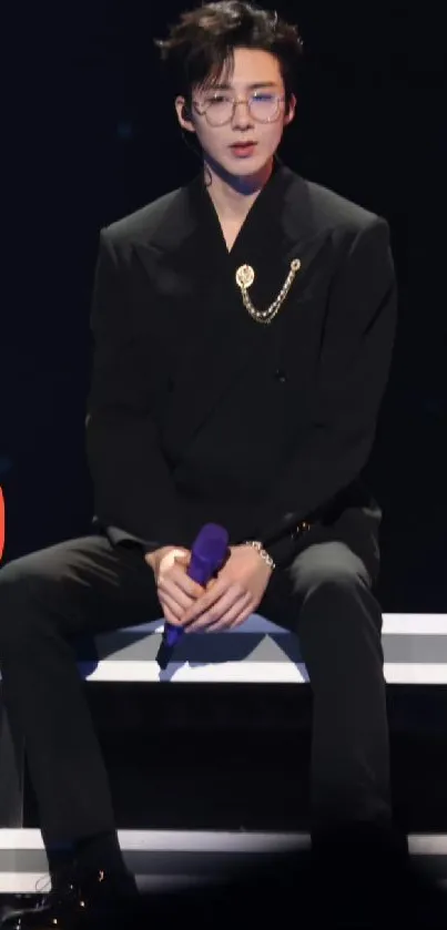 Stylish performer in black suit on stage, seated elegantly.