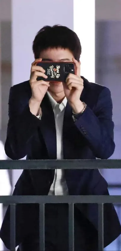 Person in dark suit holding camera on balcony.