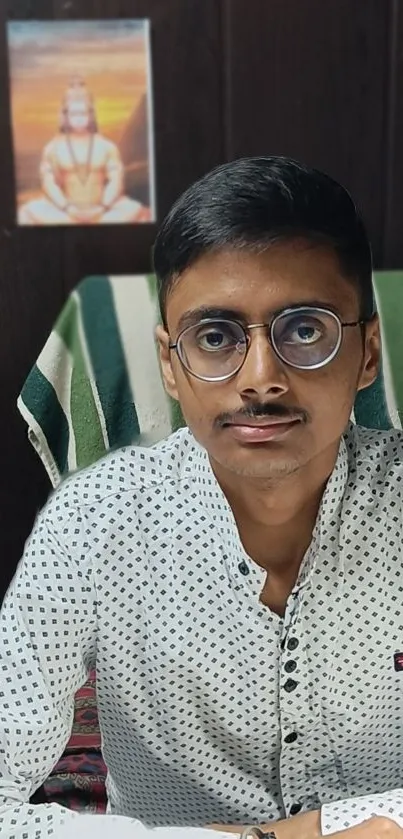 Young man with glasses sitting at a desk with a serene portrait in the background.