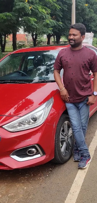 A man standing next to a red car on a tree-lined road.