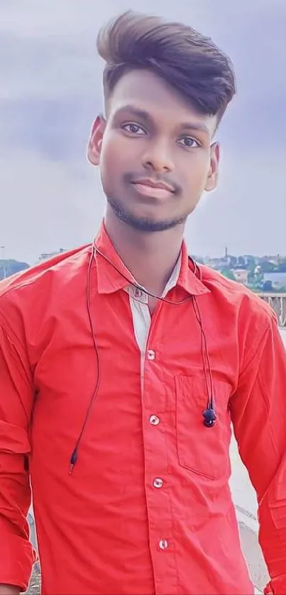 Young man in a bright red shirt standing outdoors with a cloudy sky backdrop.