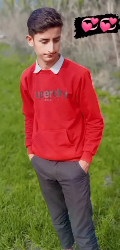 Young man in red sweatshirt stands on grassy field with heart emojis.