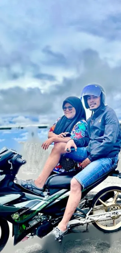 Two people on a motorcycle with a scenic cloudy background.