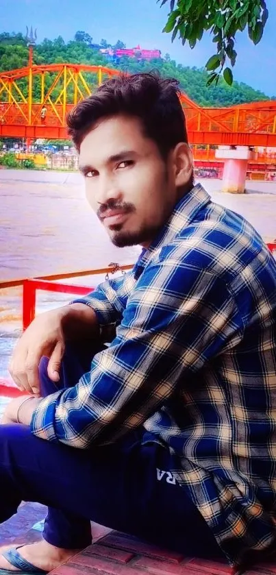 Man sitting by river with orange bridge backdrop.