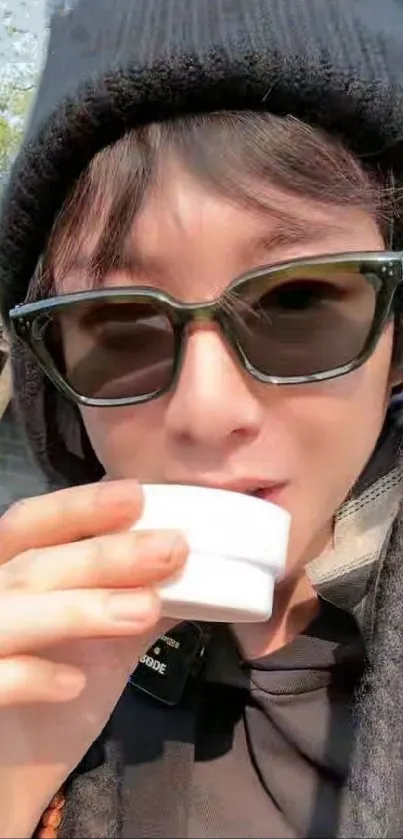 Person in sunglasses and beanie sipping from a cup outdoors.