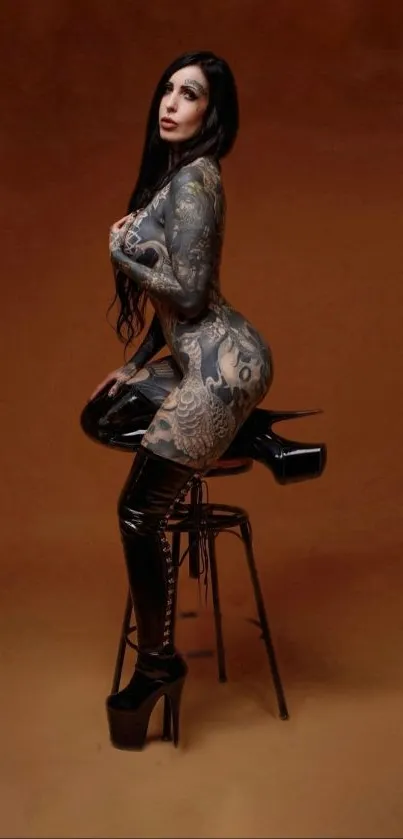 Tattooed model in black attire sitting on stool with brown background.