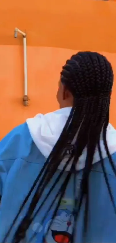 Person with braids and blue jacket in front of orange wall.