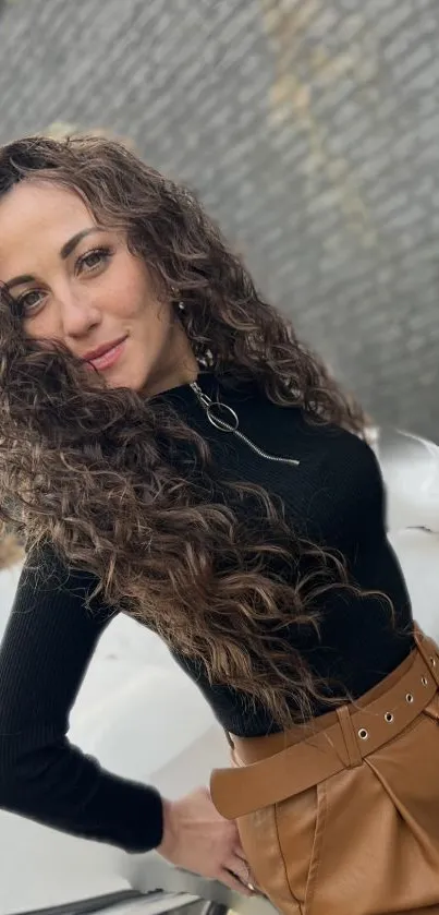 Woman with curly hair and black top poses stylishly against a brick wall.