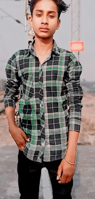 Young person in a green plaid shirt standing by railway tracks.