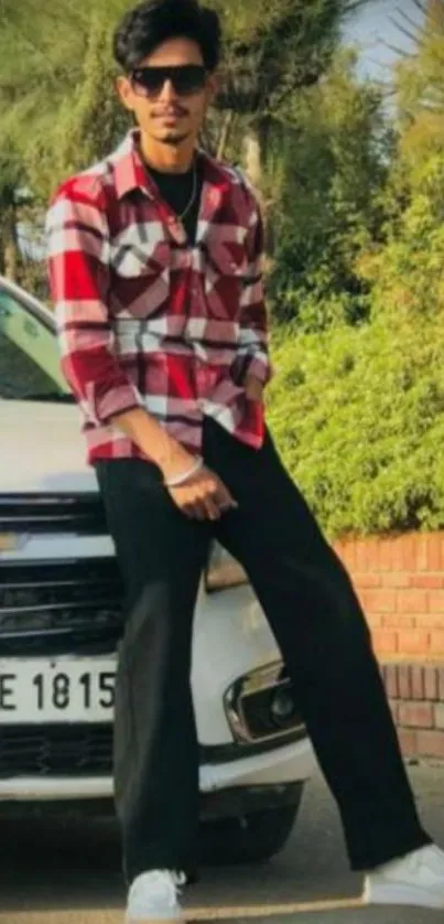 Man in red plaid shirt leaning on car outdoors.