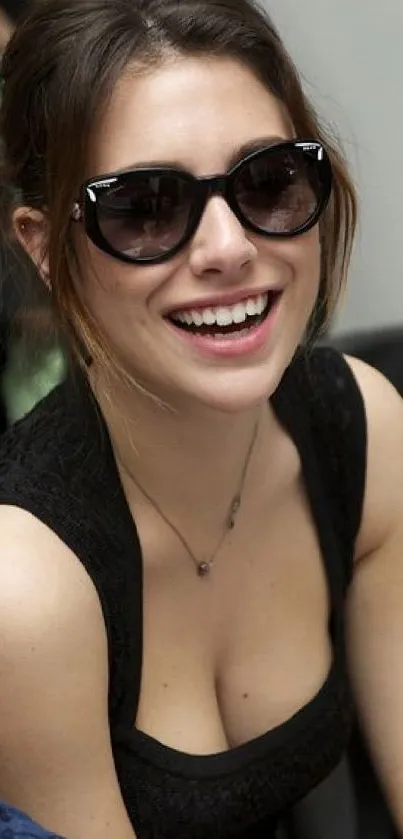 Woman in sunglasses smiling vibrantly in a stylish black outfit.