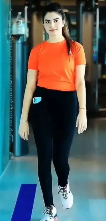 Woman in orange shirt walking in gym with mobile phone in pocket.