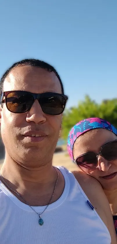 Couple taking a selfie on a sunny beach day with a bright blue sky.