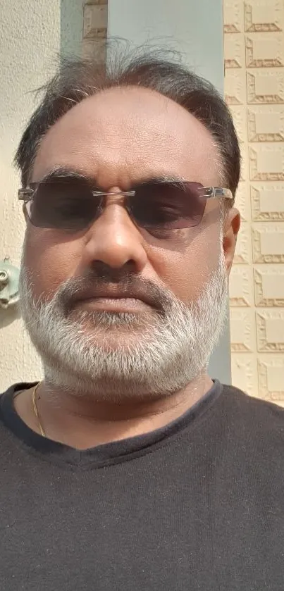 Portrait of a man in sunglasses against a textured beige wall on a sunny day.