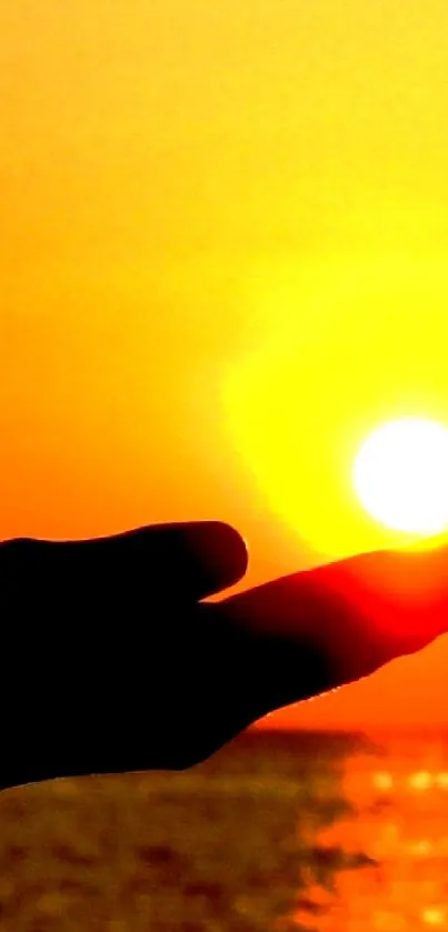 Hand silhouette holding the sun against an orange sunset over the ocean.