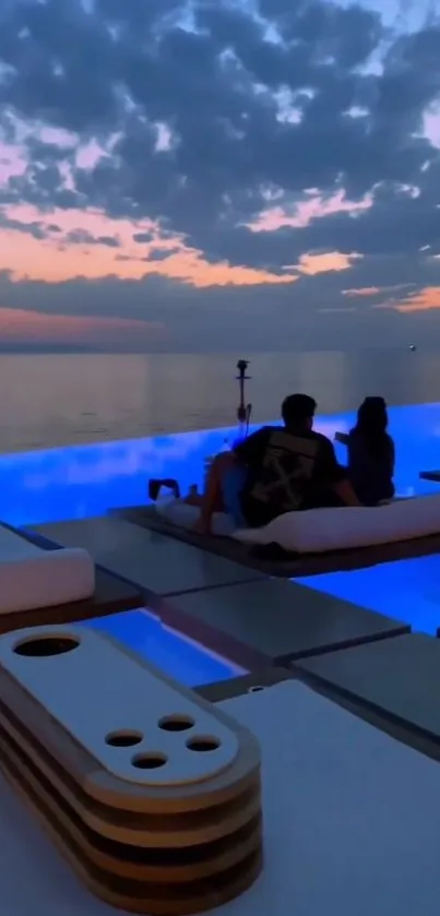 A couple relaxes by a pool under a colorful sunset sky.