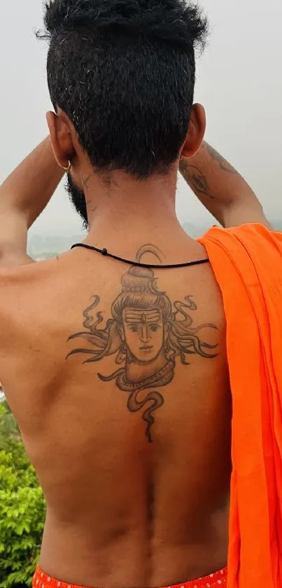 Man with Shiva tattoo stands against a natural backdrop in orange attire.