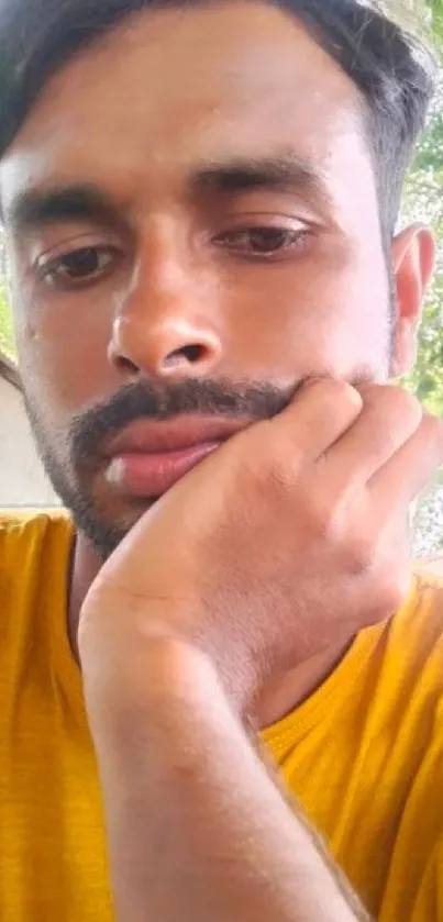 Thoughtful man in yellow shirt, greenery backdrop.